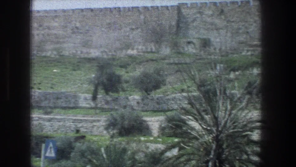 Jerusalem landscape on 8mm film, showing a crenellated wall, green terraces, small trees, palm fronds, and a roadside sign.