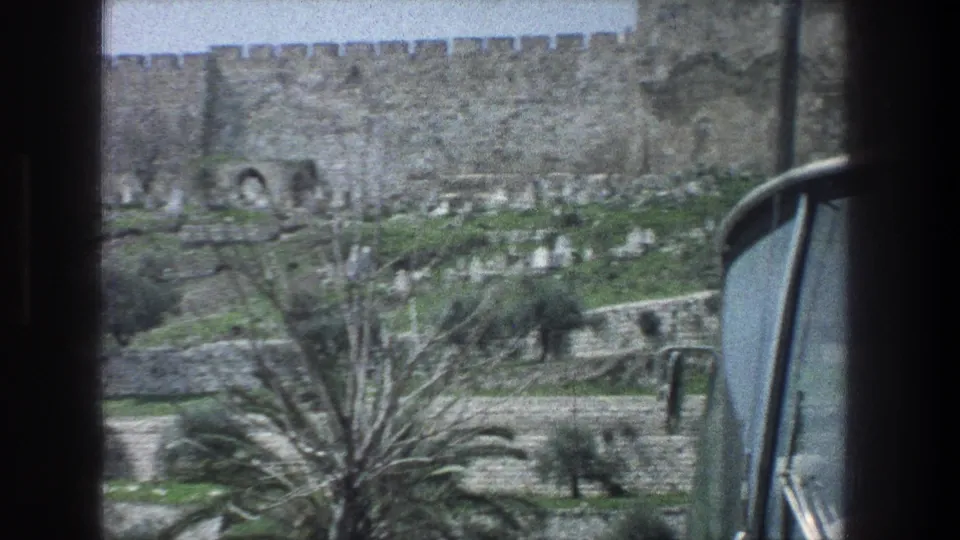 Moving-vehicle 8mm view in Jerusalem, with a car window edge, palm fronds, terraced hillside, possible cemetery markers, and stone wall.