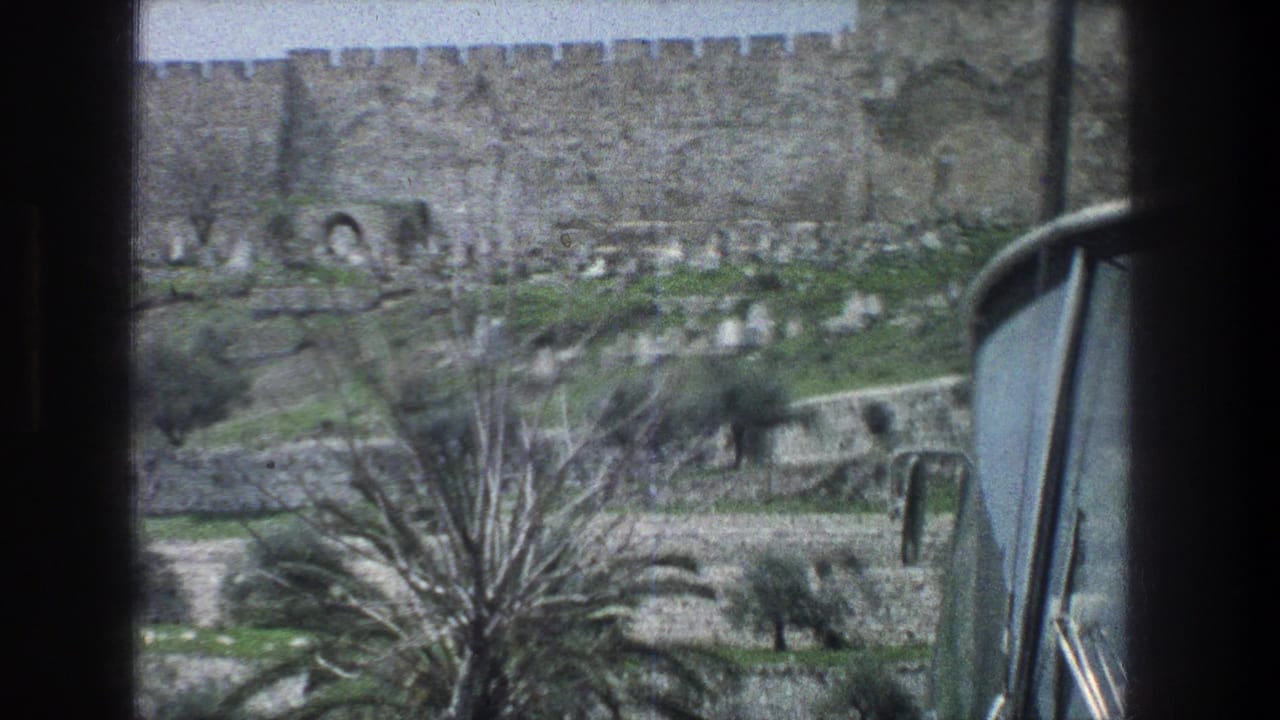 1976 Jerusalem view through a moving vehicle window, with battlemented stone wall, hillside graves, palm tree, and roadside frame edges.