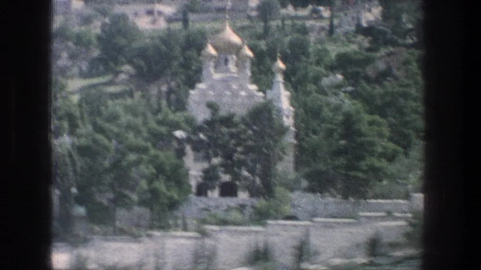 1976 Jerusalem 8mm frame showing a distant pale stone church with golden onion domes behind dense trees and terraced stone walls.