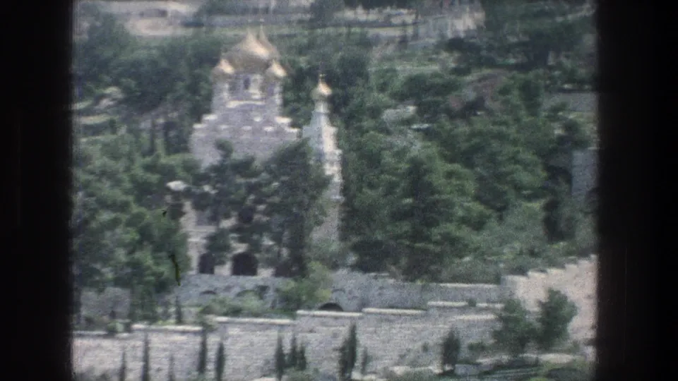 Soft 8mm Jerusalem frame from 1976 showing a golden-domed church partly obscured by trees above stone terraces and walls.