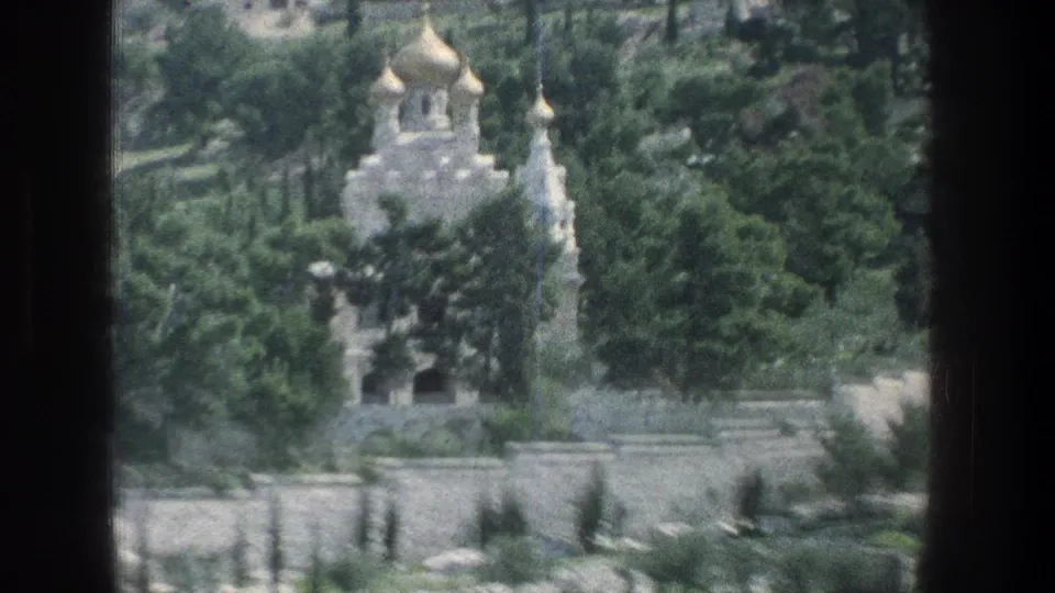 1976 Jerusalem 8mm view of a pale church with golden domes, surrounded by green trees and stone walls in bright daylight.