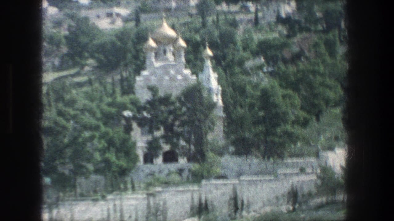 1976 Jerusalem home-movie frame showing a distant golden-domed building among trees and stone walls from a moving viewpoint.
