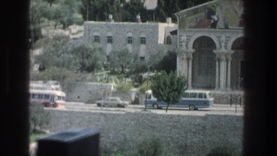 1976 Jerusalem street view with parked buses and cars in front of a stone building with arched columns and a decorated upper facade.