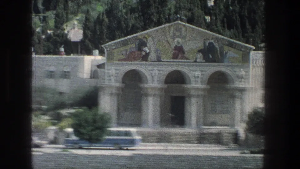 1976 Jerusalem frame showing an arched stone entrance with a mural-like upper facade, trees, road, and a parked bus in front.