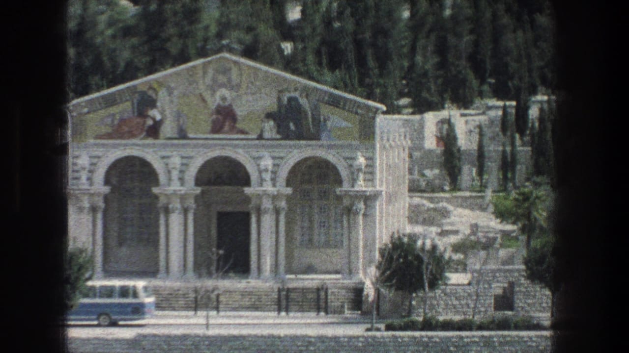 1976 Jerusalem 8mm view of a white stone building with arches, faded pediment murals, steps, cypress-like trees, and a blue vehicle passing below.