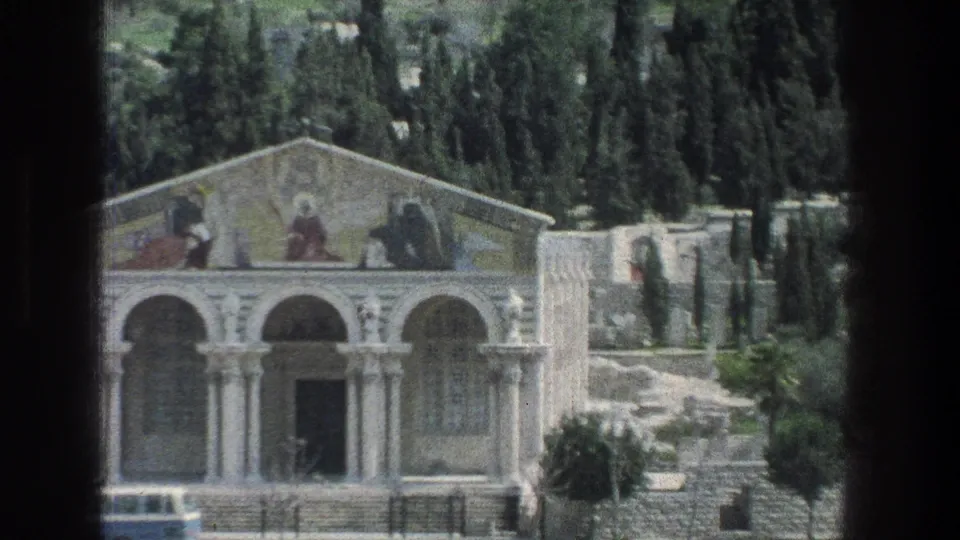 1976 Jerusalem exterior showing a white stone arched building with faded murals in the pediment, hillside trees, stone walls, and a dark central doorway.