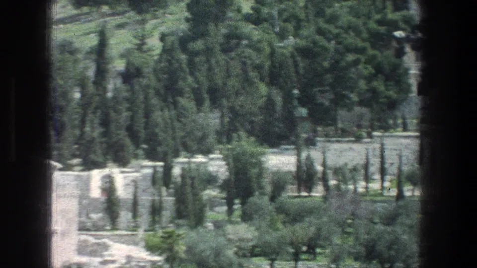 1976 Jerusalem 8mm frame of a landscaped hillside below the stone building, with cypress-like trees, pale walls, paths, and soft motion blur.