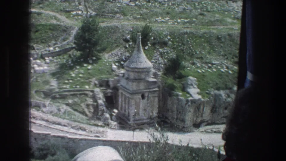 1976 Jerusalem home-movie frame showing the Tomb of Absalom, stone walls, grass, and a dark viewer silhouette in the foreground.
