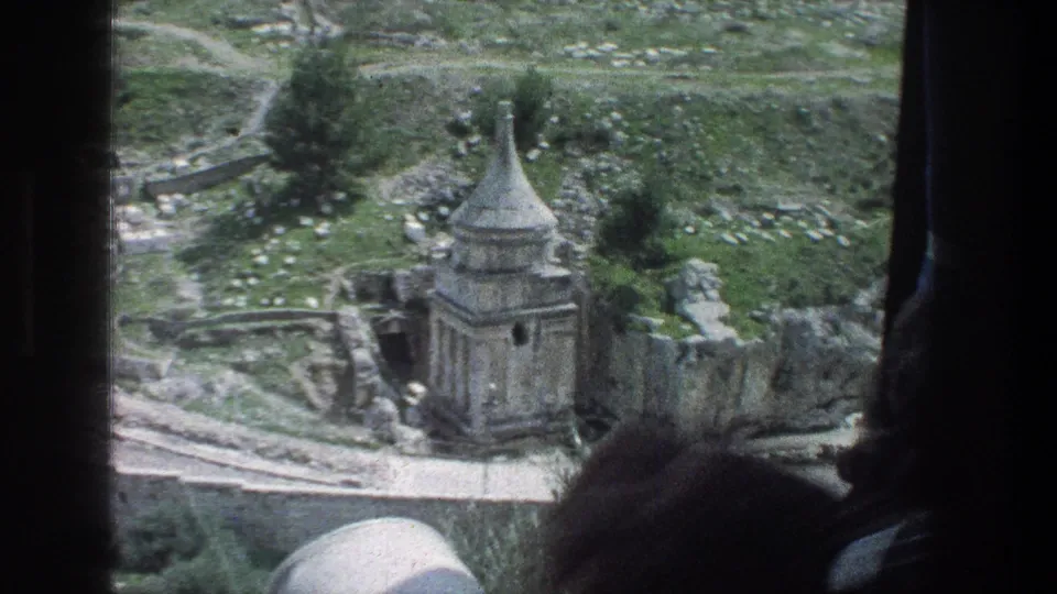 A dark passenger silhouette frames a 1976 Jerusalem view of a stone ruin, paths, grass, and scattered masonry.