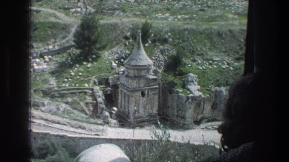 The 1976 home-movie frame looks down from a vehicle window onto a stone structure with a conical roof and surrounding hillside.
