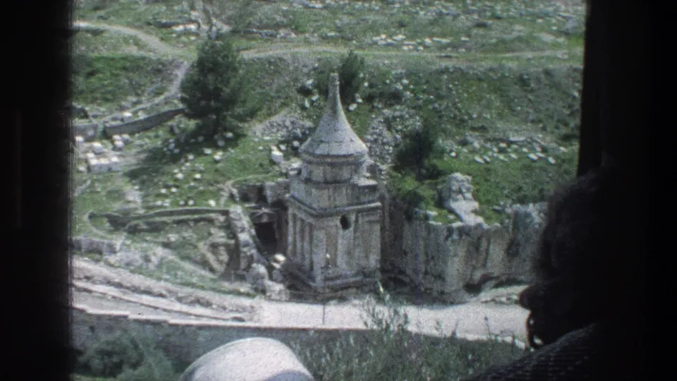 A passenger silhouette partially blocks a 1976 Jerusalem view of a stone ruin, grassy terrain, and scattered stones.
