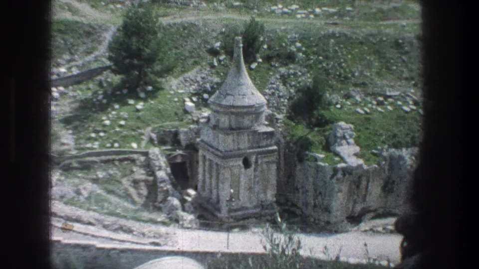 1976 8mm Jerusalem view of a conical-roofed stone structure surrounded by rocky ruin walls and patches of greenery.