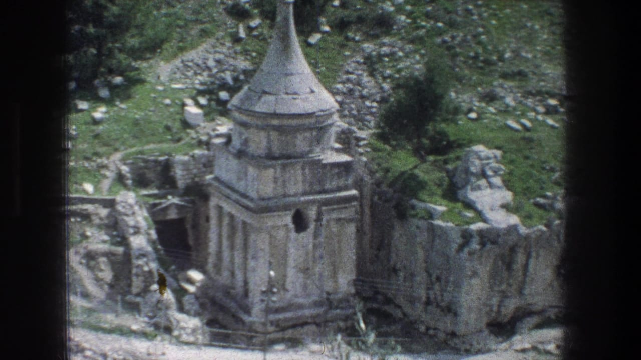 Handheld 8mm view in Jerusalem showing the stone monument from above, with rocks, low walls, and greenery around it.