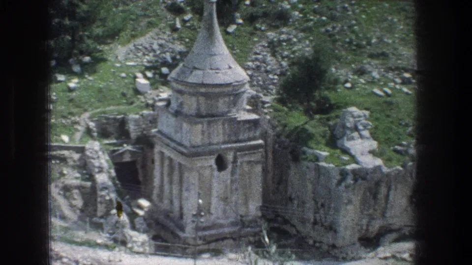 Handheld 8mm view in Jerusalem showing the stone monument from above, with rocks, low walls, and greenery around it.