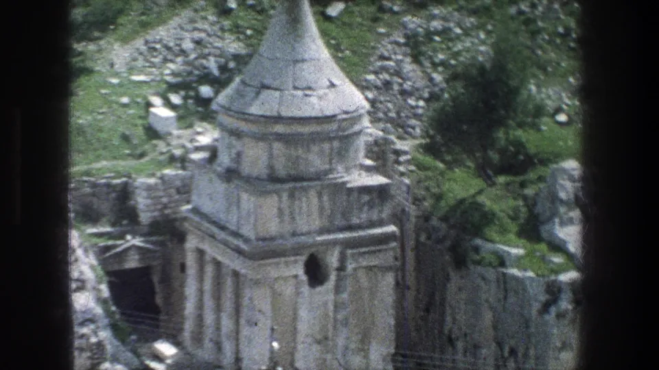 Jerusalem home-movie frame showing the conical-roofed stone monument with adjacent ruin walls, rocks, and green ground cover.