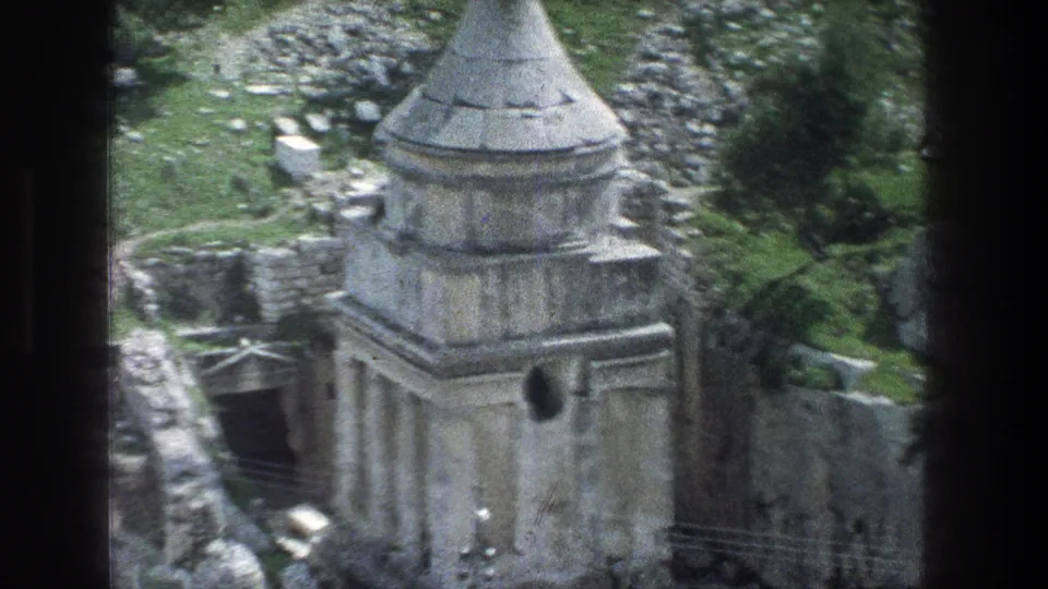 1976 Jerusalem home-movie frame showing a weathered stone structure with a conical spire, arched opening, rocks, and greenery.