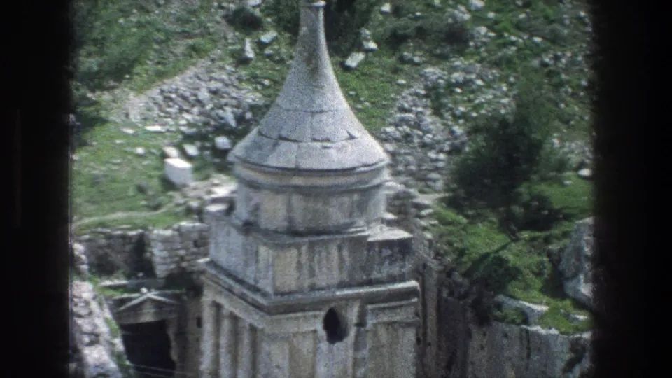 1976 Jerusalem frame showing the stone ruin from above, with a pointed spire, dark entrance, surrounding rocks, and hillside plants.