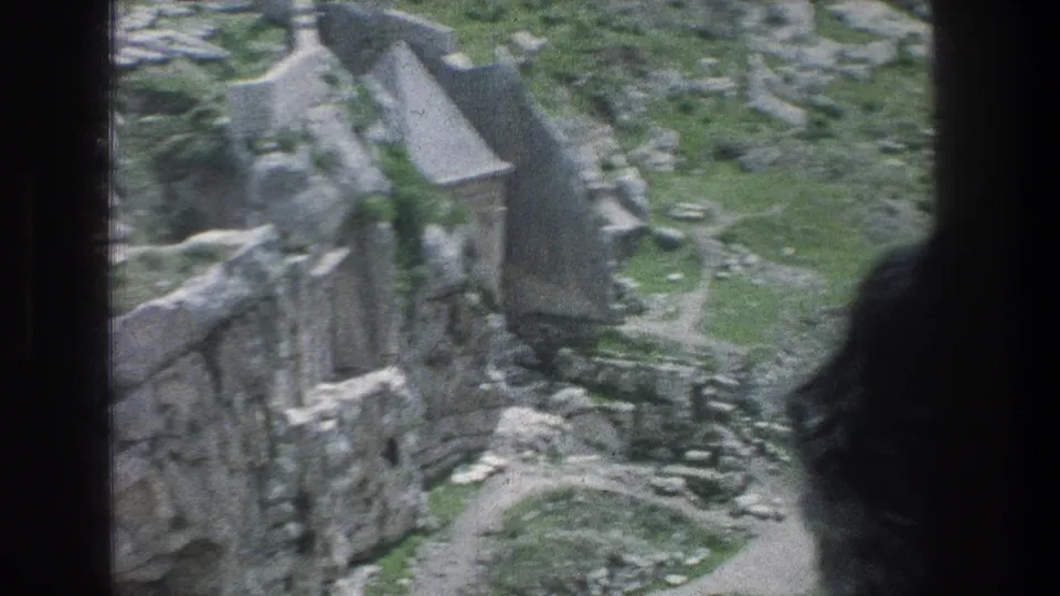 1976 8mm Jerusalem frame of cliffside ruins, gray rocks, grass, and a dark foreground edge from the shooting position.