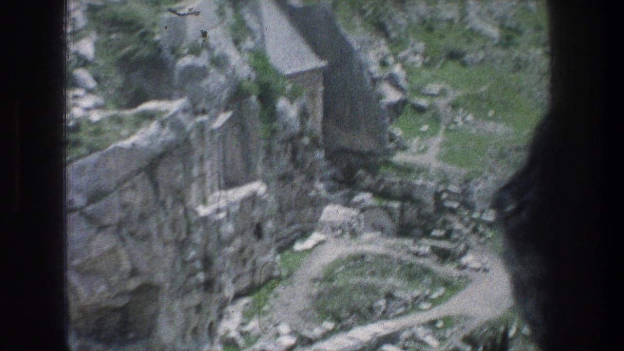 High-angle view of stone ruins, a dirt path, rocks, and greenery framed by dark vertical edges.