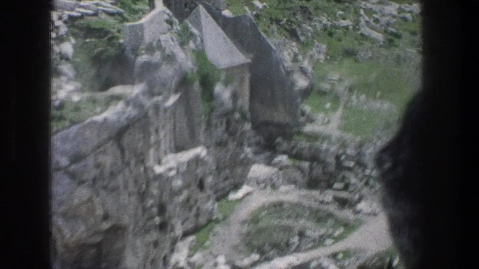 1976 Jerusalem home-movie frame with ruins, a small roofed stone structure, rocky terrain, and vegetation seen through dark foreground edges.