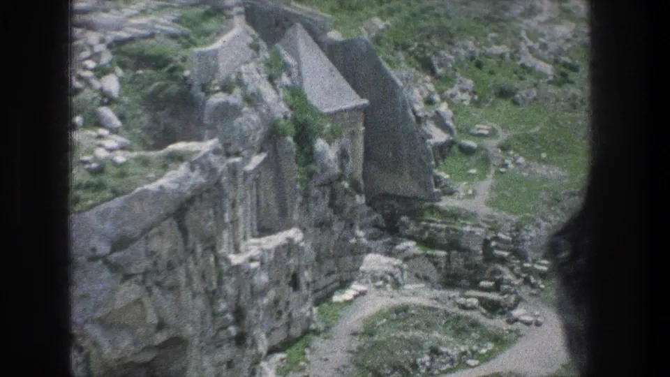 1976 8mm view of Jerusalem ruins with weathered walls, a roofed stone building, rocky ground, and a curving path below.