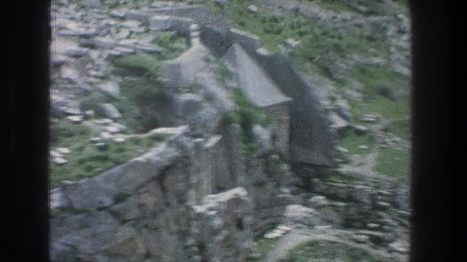 Soft 1976 Jerusalem frame showing a roofed stone structure and surrounding ruins on a rocky, green-dotted slope.