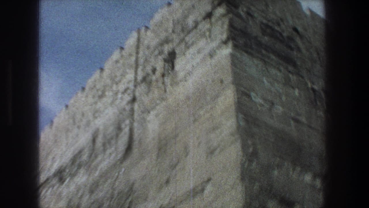 1976 8mm Jerusalem frame showing a towering weathered stone wall with crenellations near the top and pale sky behind it.