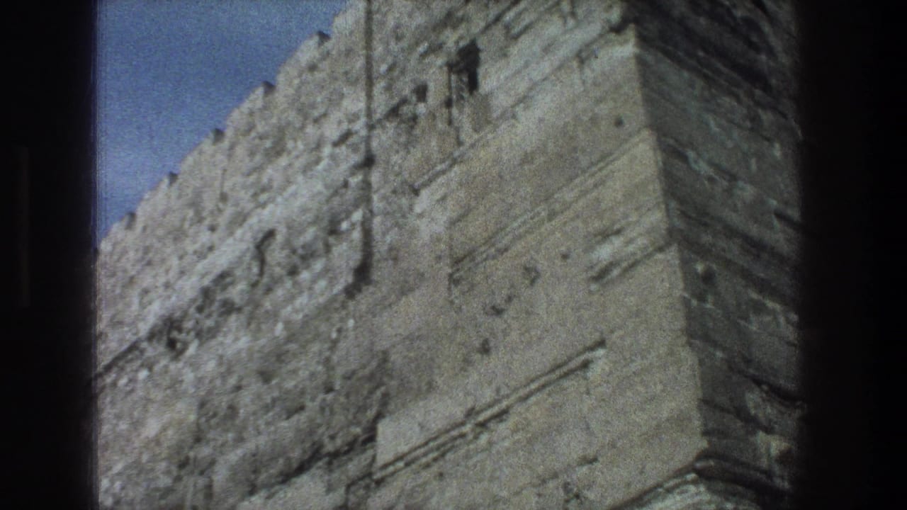 1976 8mm Jerusalem view of a stone wall corner, showing rough masonry, small dark openings, and a crenellated upper edge.