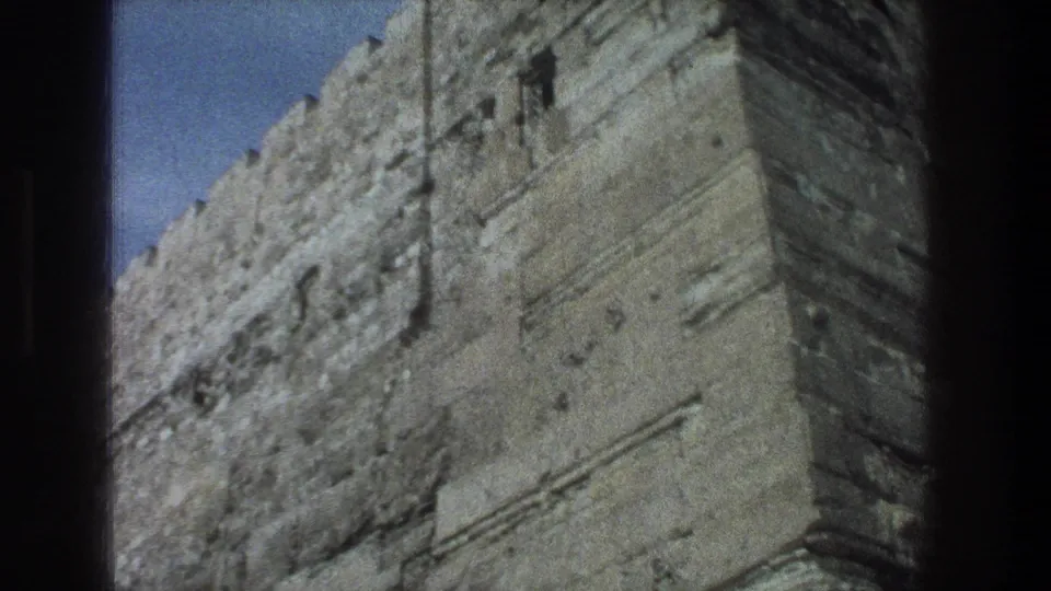 1976 8mm Jerusalem view of a stone wall corner, showing rough masonry, small dark openings, and a crenellated upper edge.