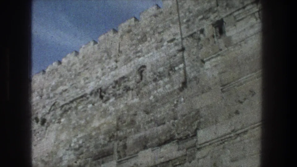 1976 8mm Jerusalem frame showing a broad section of weathered stone wall, with small openings, crenellations, and pale blue sky.