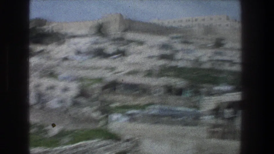 1976 Jerusalem home-movie frame with blurred stone hillside structures, pale walls, and sky during a moving pan.