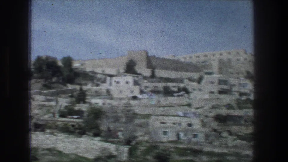 1976 Jerusalem home-movie frame showing a wider stone hillside view with terraces, scattered trees, and pale buildings.