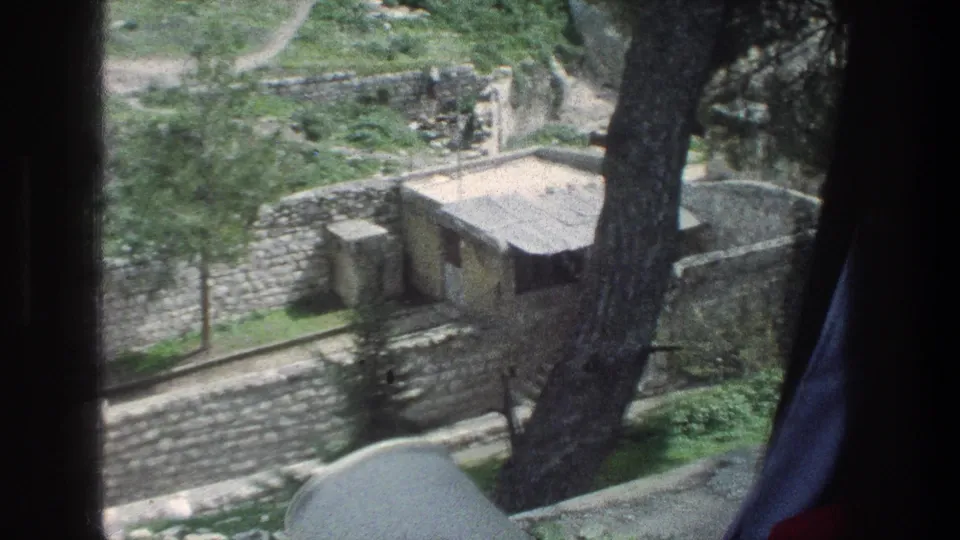 1976 Jerusalem home-movie frame with terraced stone walls, a small building, paths, and trees below an overlook.