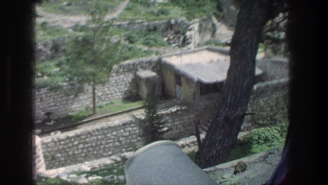 Small flat-roofed hut behind stone walls with trees and greenery in the foreground.