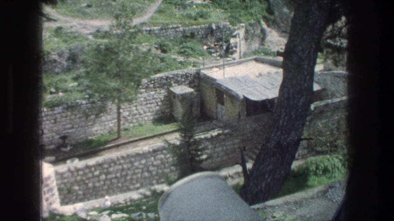 1976 Jerusalem frame with a small masonry hut, pale roof, stone walls, trees, and a path visible beyond the enclosure.