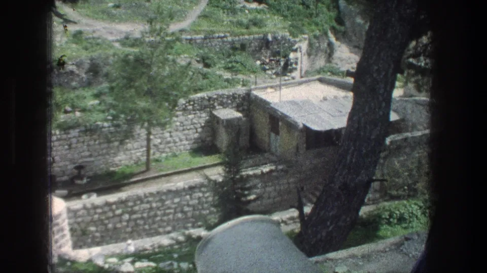 1976 Jerusalem home-movie still showing stone walls, a small hut, trees, and a curving path from an elevated, partially obscured viewpoint.