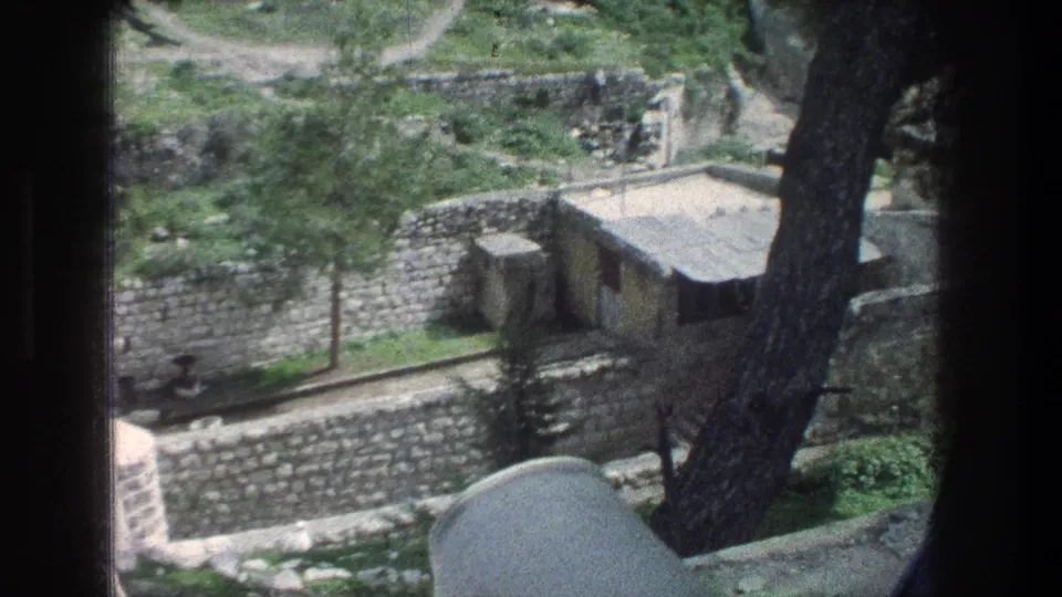 1976 Jerusalem frame with a small flat-roofed hut, stone walls, trees, and scrubby greenery, partially blocked by the foreground tree and film edge.