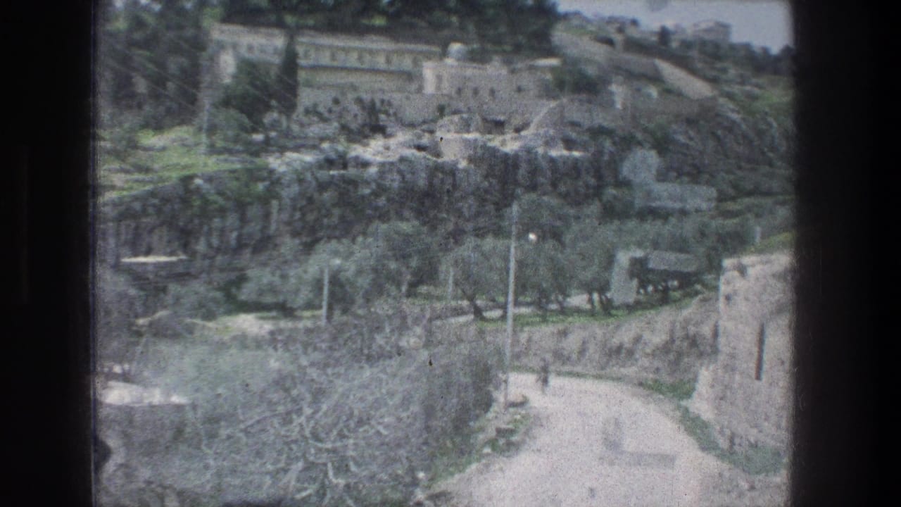 1976 8mm view in Jerusalem showing pale stone buildings, a curving dirt road, trees, and rocky hillside terrain.