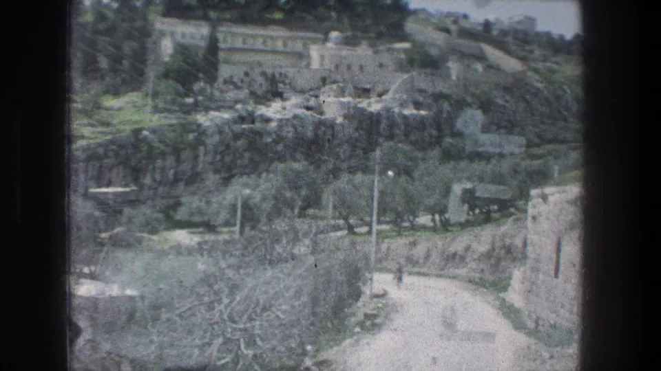 1976 8mm view in Jerusalem showing pale stone buildings, a curving dirt road, trees, and rocky hillside terrain.