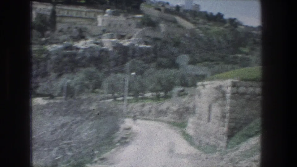 Jerusalem hillside view on 8mm film, with a pale road curving past stone walls, trees, and terraced rocky ground.