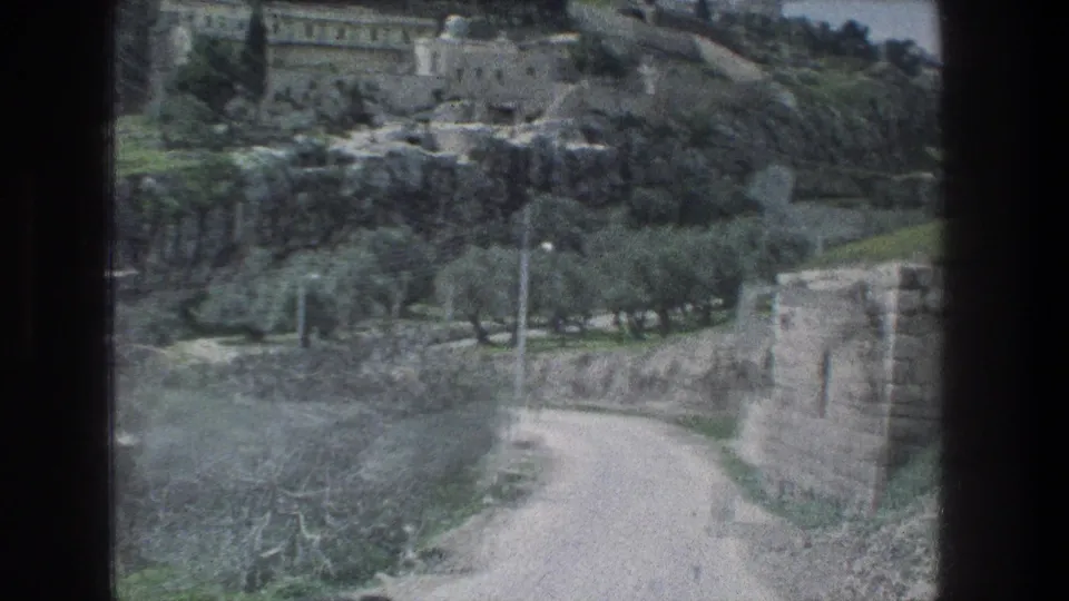 1976 home-movie frame of a Jerusalem hillside road, foreground stonework, olive trees, and pale buildings higher on the slope.