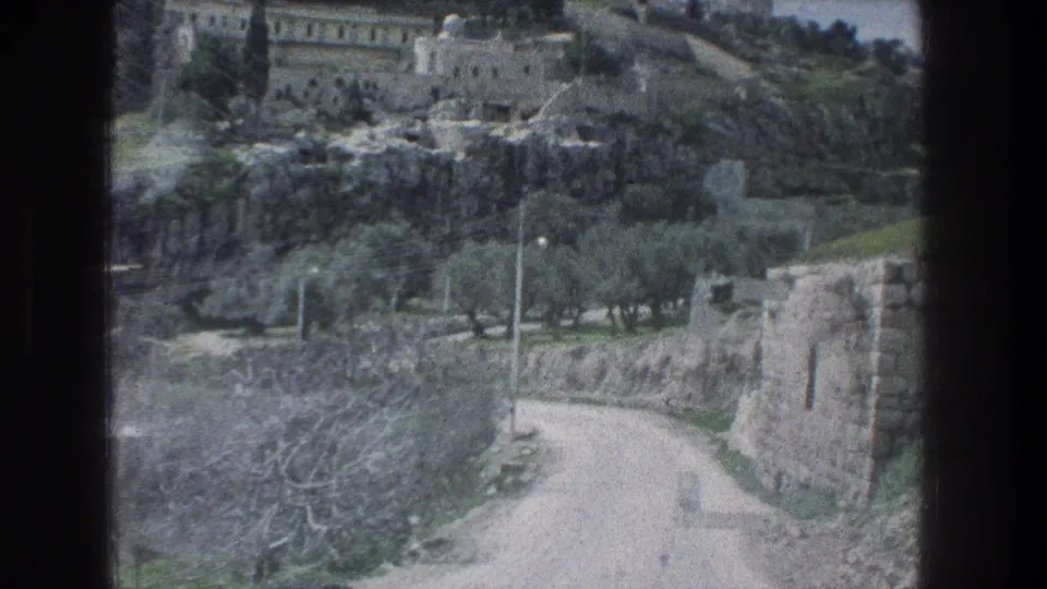 Late shot of the Jerusalem hillside view, with a curving road, stone walls, trees, rocky ground, and pale buildings in soft 8mm color.