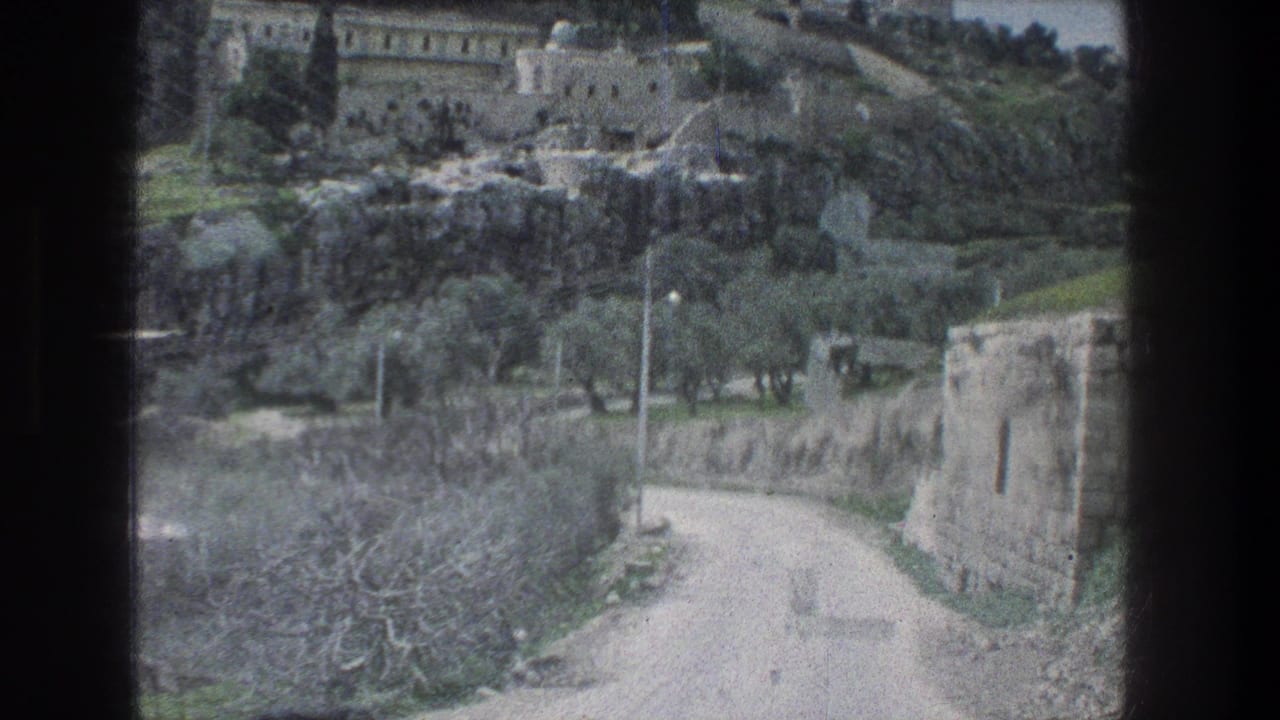 1976 Jerusalem travel footage looking from a vehicle toward a winding dirt road, stone walls, trees and hillside buildings.
