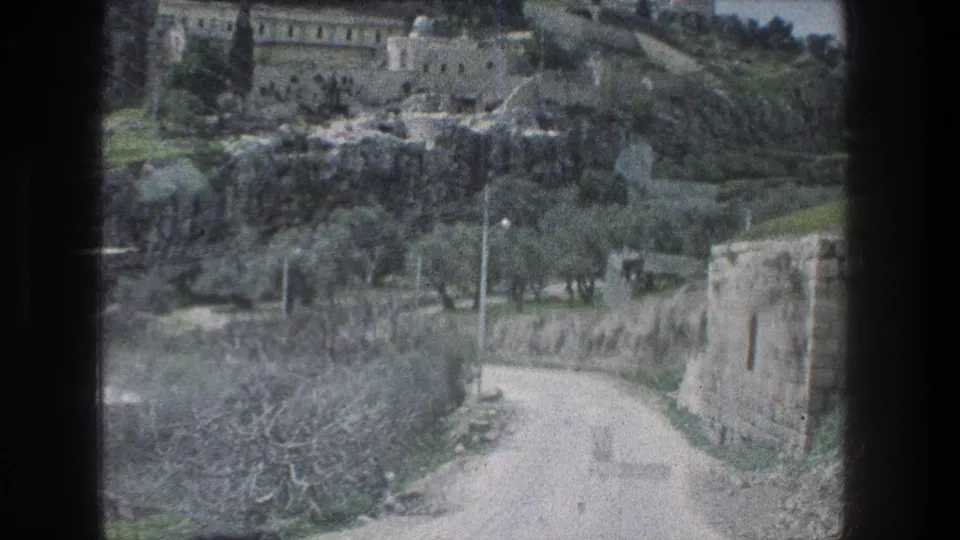 1976 Jerusalem travel footage looking from a vehicle toward a winding dirt road, stone walls, trees and hillside buildings.