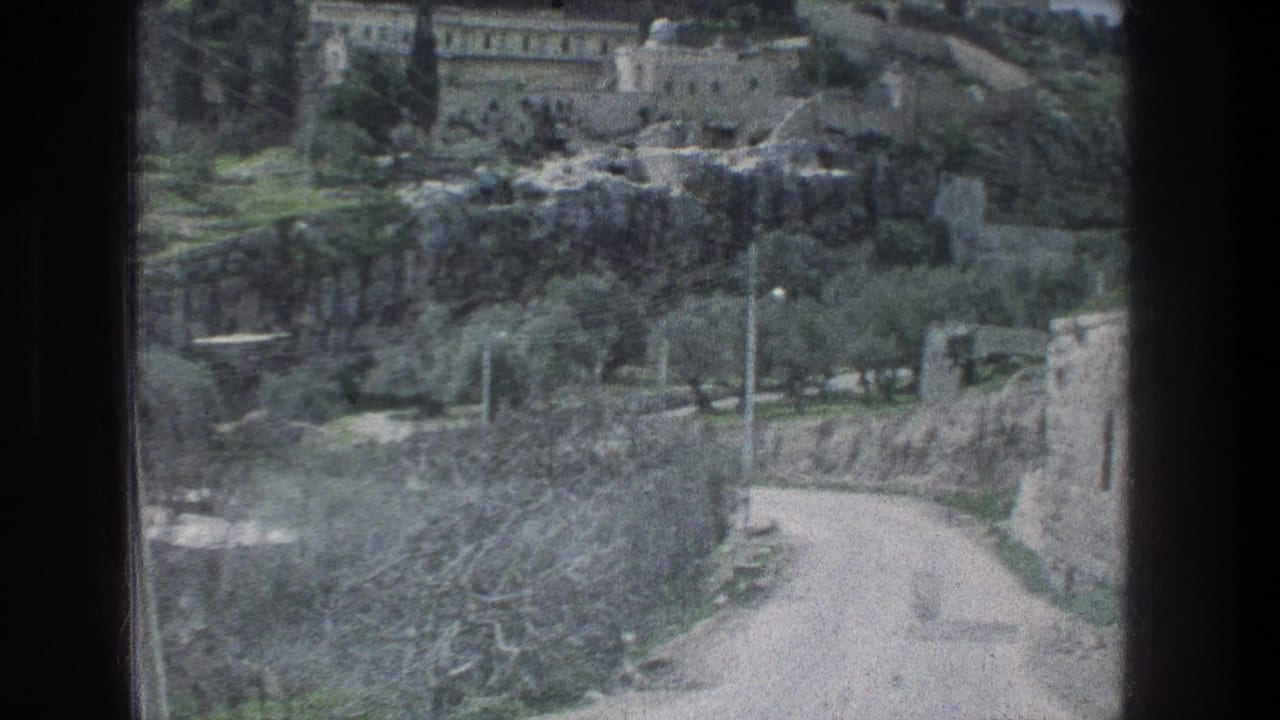 Passenger view in Jerusalem, 1976, showing a pale road beside stone walls, trees and hillside buildings through a dark vehicle frame.