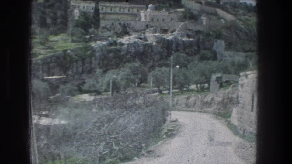 Passenger view in Jerusalem, 1976, showing a pale road beside stone walls, trees and hillside buildings through a dark vehicle frame.