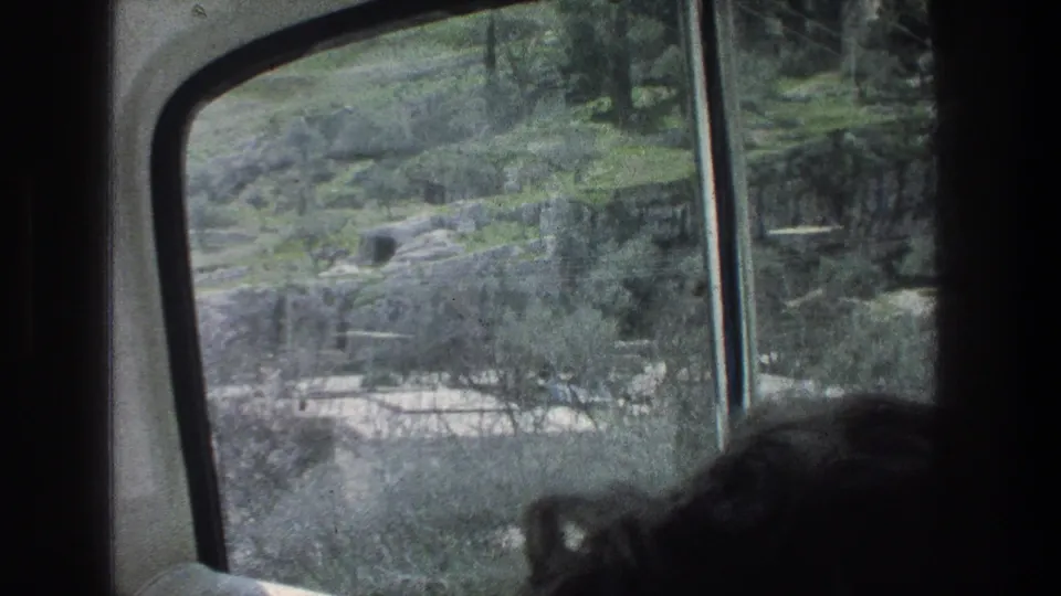 Inside a moving vehicle in Jerusalem, 1976, the camera looks past a passenger's head toward trees, stone terraces and hillside ground.