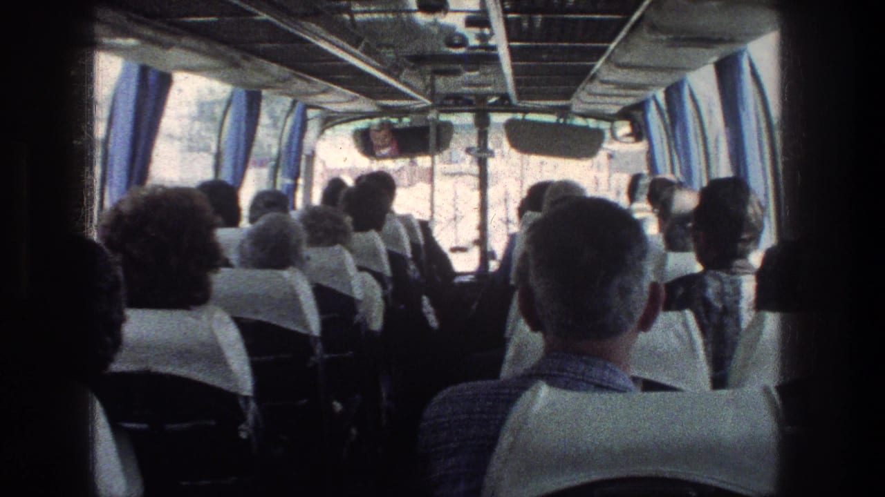 Adults seated inside a bus viewed from the rear, with curtains and overhead rails visible.