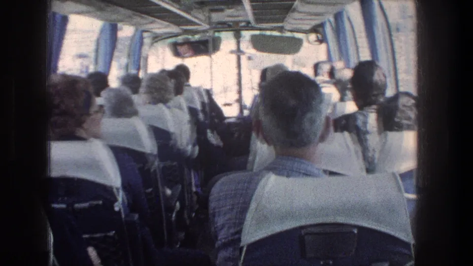 Inside a 1976 Jerusalem bus, adult passengers sit in rows beneath overhead rails as the 8mm camera looks forward from the back.